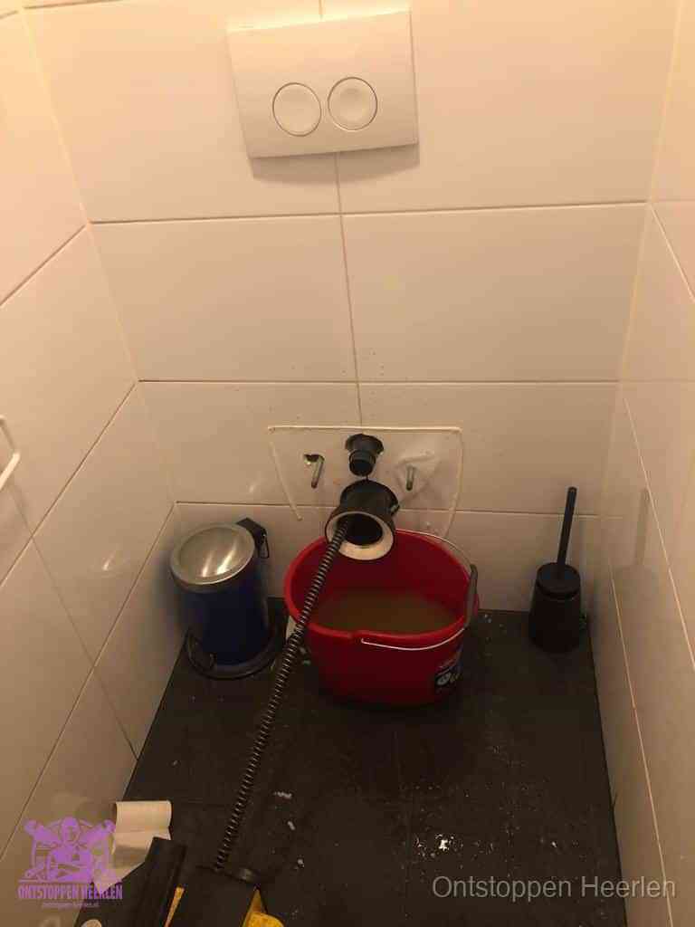 Een ontstopte wand-wc met verwijderde toiletpot waar een ontstoppingsveer in de wanduitgang verdwijnt, het achterhalen waar de verstopping zit in Heerlen