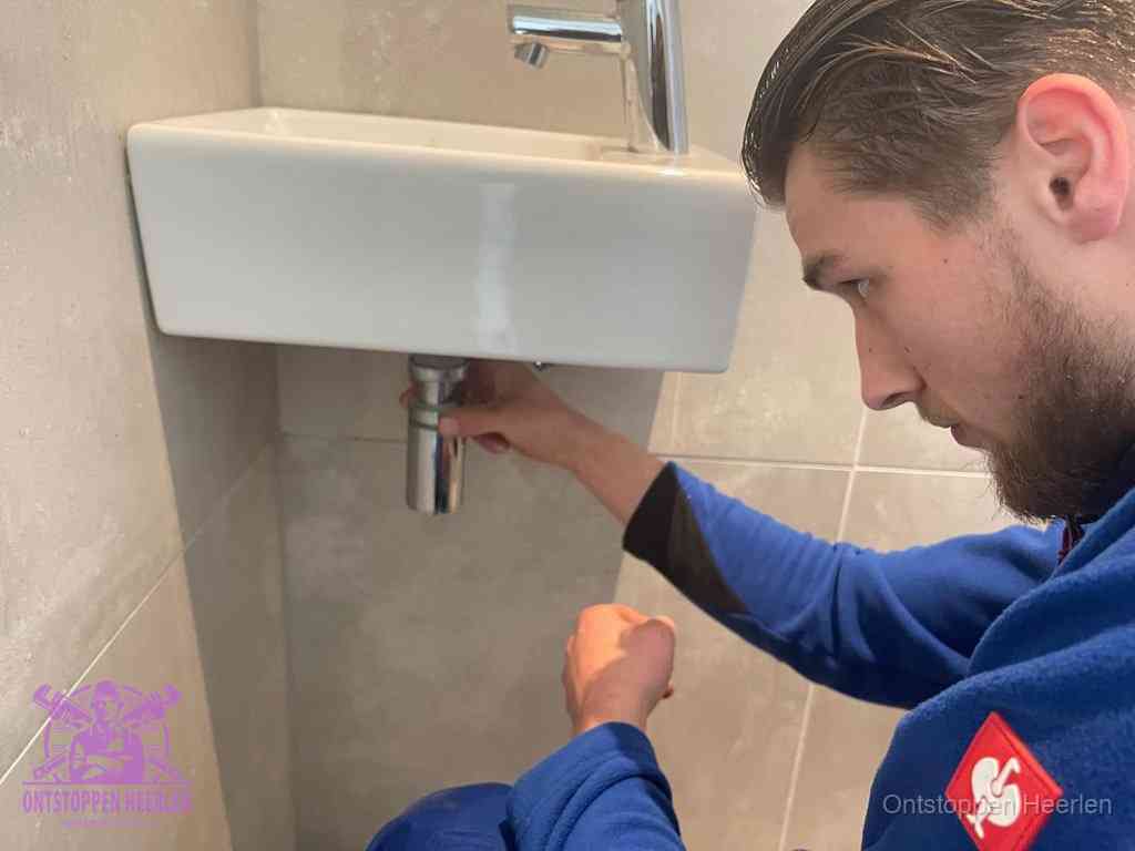 Een monteur draait met de hand de zwanenhalssifon onder een fonteinwasbak in de toiletruimte los om de bron van rioollucht in Heerlen te vinden