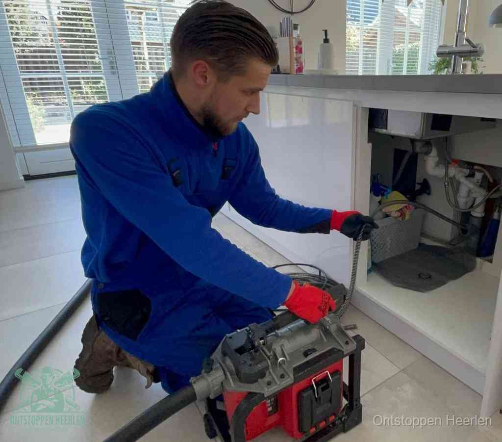 Een monteur met rode handschoenen knielt in een keuken en voert een elektrische ontstoppingsveer in tijdens het professioneel afvoer ontstoppen in Heerlen
